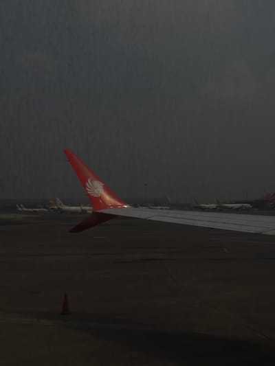 This image captures a view from an airplane window overlooking an airport tarmac in Tangerang, Indonesia. The foreground is dominated by the red and white wing of the airplane from which the photo is taken, featuring the Lion Air logo. In the background, several other aircraft are visible, some parked on the tarmac. One airplane has green accents on its wings. The environment appears to be an airport with paved surfaces and what seems to be a runway or taxiway. The sky is overcast and dark, suggesting either early morning, late evening, or inclement weather. A single orange traffic cone is visible in the lower left portion of the tarmac. The scene is devoid of any people.