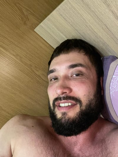 A close-up, overhead shot captures a man lying in bed, with his face taking up most of the frame. He has dark hair and a full beard, and is smiling, showing his teeth. His bare shoulder and chest are visible, with a mole on his right shoulder. The background consists of a wooden headboard and a pillow with a floral pattern. There are no visible indications of the man's location within a city or country.