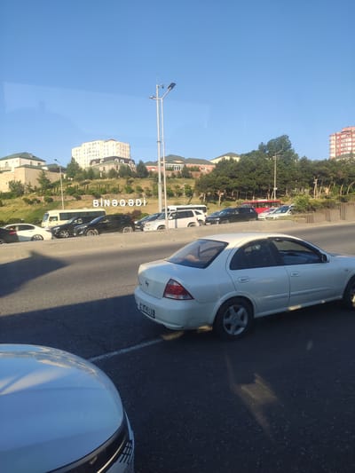 This image depicts a street scene, likely in Azerbaijan, given the "Motodrom YS" context. The photograph is taken from the perspective of someone inside a vehicle, looking out at a row of parked cars on the right side of the road. The cars are of various colors, including white, black, and red. The scene includes buildings and trees in the background, suggesting an urban or suburban environment. The sky is clear and blue, indicating a daytime setting with good weather.

The image captures a static moment, with the cars parked along the roadside. There are no people clearly visible in the foreground or midground. The overall impression is one of a typical street scene, possibly during a pause in traffic or a moment where vehicles are parked for an extended period.

A notable detail is the text visible on the license plate of one of the white cars: "90-HH-136". Another piece of text, appearing vertically on the left side of the image and partially obscured by reflections, seems to be a place name or a sign, reading "BİNƏQƏDİ". The light suggests it's daytime, likely late afternoon or early morning due to the angle of the shadows. The reflection on the left suggests the photograph was taken through a window, possibly of a bus or car.