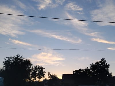 The image depicts a dusky sky over what appears to be a rural or semi-rural area in Cîrpești, Moldova. Two dark power lines stretch horizontally across the frame, one higher and one lower, creating a division of the sky. Wispy, light-colored clouds are scattered across the pale blue sky, with hints of yellow and orange near the horizon, suggesting either sunrise or sunset. Silhouetted against the sky are the dark shapes of trees and the rooftops of several buildings, indicating a settlement. The overall scene is one of quiet twilight.