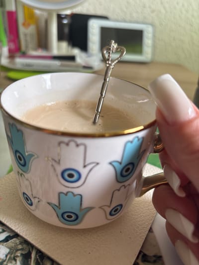 A close-up, slightly tilted shot shows a hand holding a partially filled white coffee cup with blue Hamsa hand designs and a gold rim. A small silver stirring spoon with a decorative handle sits inside the cup. The hand has long, white, squared-off artificial nails. The cup rests on a light-colored surface, possibly a coaster. In the blurred background, there are various objects including what appears to be a small white-framed mirror or picture, tubes, and other items suggesting a desk or table setting. The location of Werl, Germany cannot be determined from the image provided.