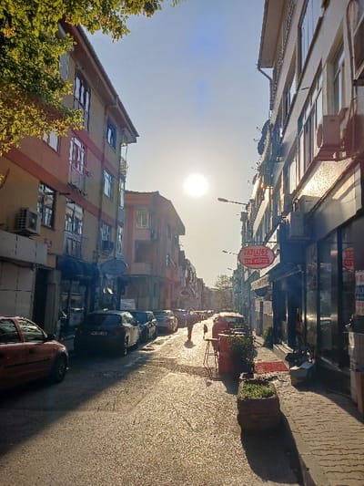 The image shows a narrow street in Istanbul, Türkiye, located at 40.962 latitude and 28.825 longitude. The street is lined with buildings on both sides, with cars parked along the edge of the street. The street is paved with cobblestones and there are a few potted plants along the side. The sun is shining brightly in the sky, casting a warm glow over the scene. A single person walks down the middle of the street, heading towards the sun. To the left, a red car is parked near the beginning of the street, facing the opposite direction of traffic. In the center of the image are several parked cars, some in front of shops and others at the curb. To the right, the image features several buildings that are all multi-story. There is a sign in the window of one building that reads "ÇATI CAFE." The sign is in Turkish and refers to the roof of the building.