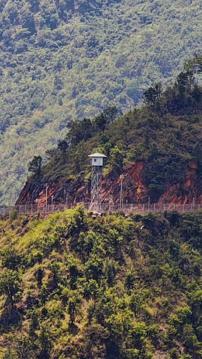 The image shows a lookout tower in Lũng Cú Commune, Vietnam. The tower is a small, white structure with a metal frame, situated on a hillside. A fence runs along the hillside near the tower. The surrounding environment consists of lush, green vegetation and steep terrain. The scene appears to be outdoors and is captured during daylight hours. The image appears to be taken from a distance.