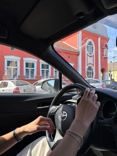 View from inside a car, focused on the dashboard, steering wheel, and the driver's hands, presumably driving, partially revealing the exterior view of a building with red facade and white architectural details. The building has large oval windows and what appears to be a sculpture or architectural feature on the roof. Through the car windows and side mirror, additional vehicles and parts of another building can be seen. The car license plate visible in the side mirror suggests the location might be in Serbia, specifically RU017-AH. Based on the architectural style and location indicated by the license plate, it is likely the building in view is the Gradska kuća u Rumi (Ruma Town Hall), located in the city of Ruma, Ruma Municipality, Serbia.