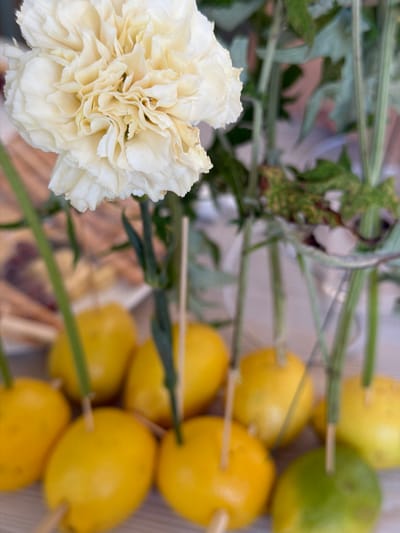 A close-up, slightly blurred photograph captures a still life arrangement featuring a creamy white carnation flower and several small lemons on skewers. The carnation, positioned in the upper left quadrant, displays intricate, ruffled petals with subtle yellow undertones. Behind it, the stems of other flowers or greenery are visible, along with a blurred background suggesting an indoor setting with some light wooden elements. The lemons, arranged in the lower half of the image, are round and appear to be a mix of yellow and one green, with thin wooden skewers inserted into them. The lighting is soft, creating a gentle ambiance. The image does not contain any people, actions, or interactions. The provided context of Heijen, Netherlands is not visually represented in the image itself.