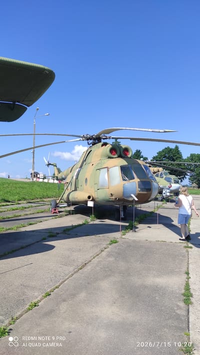 An outdoor, ground-level, full-shot photograph captures two camouflaged helicopters on a sunny day in what appears to be a museum or display area. The primary focus is on the larger helicopter in the foreground, a military-style aircraft with a tandem rotor system. Its camouflage is a mix of tan, green, and brown. The helicopter is parked on a concrete surface, with grass growing in the cracks. A red support is visible under one of its wheels. In the background, a second, smaller helicopter of similar camouflage is visible.
A woman with blonde hair, wearing a white t-shirt and blue shorts, is standing near the front of the larger helicopter, facing away from the camera, and looking towards the aircraft. She is holding a white object in her right hand. The sky is a clear, bright blue with a few wispy clouds. A street lamp and a green grassy hill are visible behind the helicopters, with some buildings and trees in the distance. The watermarks "REDMI NOTE 9 PRO" and "AI QUAD CAMERA" are present in the bottom left corner, and a date stamp "2026/7/15 10:20" is in the bottom right corner. The location is identified as Лашанскі сельскі Савет, Belarus.