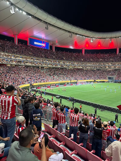This is a high-angle view inside a stadium, likely during a soccer game, located in Zapopan, Mexico, as evidenced by the advertisements in Spanish. The stadium is densely packed with spectators, many wearing red and white striped shirts, possibly indicating support for the same team. In the foreground, fans are seated in red stadium seats, some holding up their phones to capture the action. In the distance, the soccer field is visible, with players engaged in a match. A large video board displaying "!GOOOOOL!" hangs from the stadium roof, which is illuminated with red lights. Numerous advertisements are placed around the field. The sky is dark, indicating the game is being played at night.