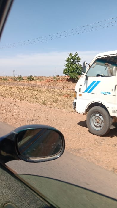 The scene depicts an outdoor environment in Barakin Columbai, Nigeria, captured from the interior perspective of a moving or stationary vehicle. In the foreground on the left, a dark-colored exterior rearview mirror reflects a portion of the road and a blurred image. Below the mirror, a section of the vehicle's window and textured door panel is visible. To the right, a white van, identified by blue diagonal stripe decals and "Mazda" branding on its side, is positioned on an unpaved, reddish-brown dirt shoulder adjacent to a paved road. The van's front passenger side, including its headlight and a dark-colored wheel with a silver hub, is visible. No individuals are discernible within or around the vehicles. The background features arid, sandy ground with sparse, low-lying vegetation and small bushes. A single green-leafed tree stands behind the white van. The sky is clear and blue, extending across the upper portion of the frame, with thin, dotted power lines or communication lines traversing horizontally. The lighting suggests daytime conditions.