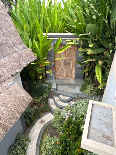 The image shows a view from above of an outdoor area with lush tropical vegetation. In the foreground, the thatched roof of a building is visible, partially obscuring a grey wall. A curved pathway made of light-colored pebbles and dark grey edging winds through the greenery, with decorative floral patterns embedded in the pathway. Small green bushes and flowering plants with orange and yellow blossoms are situated along the path.

Further into the scene, a set of grey stone steps leads up to a weathered wooden double door set within a dark stone wall. Behind the door and surrounding the steps, large, vibrant green leaves of tropical plants create a dense backdrop. Some red and orange flower clusters are visible among the foliage.

To the right of the steps, a concrete planter or ledge with a rough, weathered surface is positioned at an angle. The overall impression is of a serene, tropical garden setting, possibly part of a resort or private residence in Abang, Indonesia, given the architectural style and vegetation. There are no people visible in the image. The lighting suggests it is daytime, with natural light illuminating the scene.