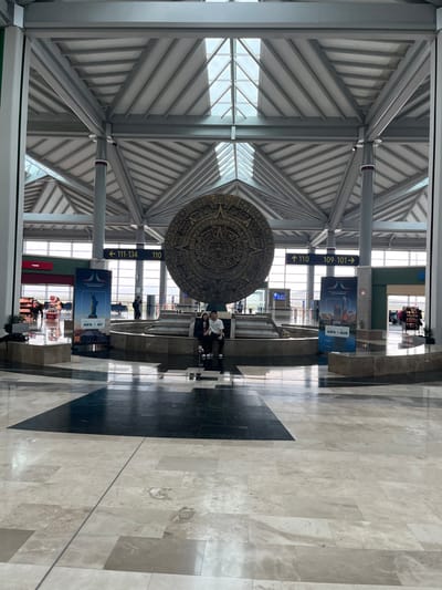 The image displays the interior of a modern airport terminal at Felipe Ángeles International Airport (AIFA) in San Miguel Jaltocan, Mexico. A large, circular, dark-colored relief sculpture, resembling the Aztec Sun Stone, is prominently positioned in the center, set within a multi-tiered, light-colored stone base that appears to be a water feature or planter. Two individuals are seated on the edge of this stone base, facing the camera. One person on the left wears dark clothing, while the person on the right wears a white top and dark trousers. The terminal features a high, vaulted ceiling with exposed metallic structural beams and multiple skylights. The floor consists of polished stone or marble tiles laid in geometric patterns using shades of beige, grey, and black. Overhead signage with yellow arrows indicates gate numbers: "← 111-134", "↑ 110", and "→ 109-101". Two freestanding banners are visible; one on the left shows the Statue of Liberty with "AIFA + NY", and another on the right displays a cityscape with "AIFA + AUS". Multiple indistinct figures are present in the background, moving throughout the spacious area.