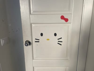 The image shows a close-up of a white door adorned with Hello Kitty decals. A red bow is placed on the upper part of the door, and the iconic Hello Kitty face, complete with black eyes, a yellow nose, and black whiskers, is centered on a square panel. The door has visible paneling and a white painted finish. To the left of the Hello Kitty face, a silver doorknob is visible, suggesting this is a functional door. Next to the doorknob, on an adjacent surface which appears to be a wall or another door, there is a light switch. The lighting suggests an indoor setting, possibly during the daytime given the overall brightness. The background is mostly out of focus, but hints at a room. The overall impression is one of a personalized and perhaps child-friendly space. The location context provided, Мухавецкі сельскі Савет, Belarus, indicates a rural or suburban area in Belarus.