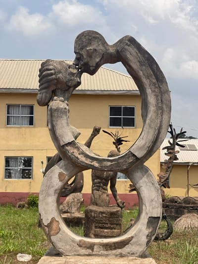 The image depicts an outdoor scene in Umunekwu, Nigeria. The primary subject is a large, weathered concrete sculpture positioned centrally, featuring two stylized, emaciated human figures intertwined in an embrace and kiss, forming a continuous, roughly figure-eight or double-loop structure. The upper figure leans over to meet the upturned face of the lower figure, whose head is held by the upper figure's arm. The sculpture exhibits significant material degradation and flaking, particularly on its lower sections.
In the background, a two-story building with a light yellow-cream facade and a beige corrugated metal roof is visible. It features multiple rectangular windows with white frames. To the right of the central sculpture, another concrete sculpture of a male figure with a raised arm and distinct musculature stands on a pedestal, which bears carved text including "OKORIE," "UCHECHUKWU," and a numeric sequence. Further abstract, spiky sculptures are visible in the mid-ground. The immediate foreground and surrounding areas consist of green grass under a partly cloudy sky.