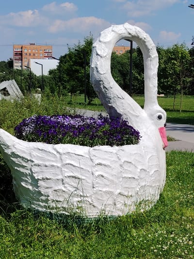 A large, white, textured swan-shaped planter filled with purple flowers sits on a green grassy lawn. The swan has a long, curved neck and is positioned in the foreground, occupying most of the frame. In the background, there are trees, a paved walkway, streetlights, and a multi-story brick building. The sky is blue with scattered white clouds. The scene appears to be in a park or public outdoor space in Dolgoprudny, Russia.