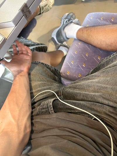 The image shows a close-up view of a person's legs and arm inside what appears to be public transportation, possibly a train, given the seating and tray table visible. The person is wearing grey jeans and white socks with athletic shoes. A white cable is visible running across the jeans. The background shows muted colors and the environment suggests an indoor setting. The image does not contain any specific elements that would indicate the location is Rivesaltes, France.