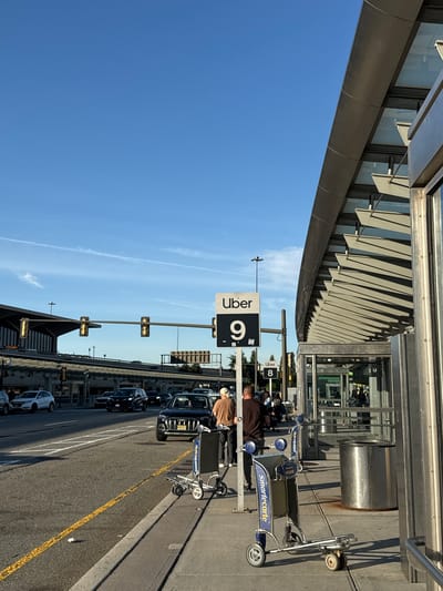 The image depicts a bustling outdoor scene at what appears to be Newark Liberty International Airport, under a bright, clear blue sky with a few wispy clouds, suggesting sunny weather during the late morning or afternoon.
On the left, a modern building facade, likely part of an airport terminal, features metallic horizontal panels and angled sun shades. Adjacent to the building, a paved roadway or drive lane is visible, lined with cars, suggesting a pickup or drop-off area. A black SUV is prominently featured among the vehicles.
Several people are present, mostly appearing to be waiting or moving luggage. One person in a brown jacket stands near a luggage cart, and another in a tan shirt is partially visible behind them.
Key objects include multiple blue and silver SmartCarte luggage carts, some clearly branded with "smartecarte" and "NEWARK." A prominent black sign with white lettering displays "Uber 9," indicating a designated ride-share pickup zone. Further down the road, another white sign with black text reads "Uber 8." Street lights or traffic signals are mounted on poles along the street. The road surface has distinct yellow and white markings.
The scene conveys a typical, functional airport environment, with cues like the "Uber" signs and "NEWARK" on the carts strongly indicating its location in Newark, United States of America.