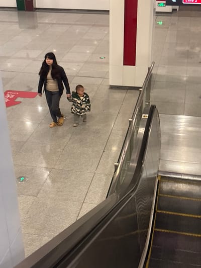 A woman with long black hair wearing a black jacket and grey pants walks on a grey tiled floor, alongside a small child wearing a camouflage jacket and grey pants. They are walking away from a red directional sign. An escalator is partially visible in the upper right corner. The scene is lit by overhead lighting, and the perspective is from above and at an angle.