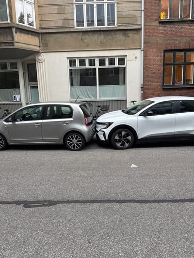 Two cars are parked bumper to bumper on a street in Frederiksberg, Denmark. The car on the left is a silver Volkswagen Up, and the car on the right is a white electric vehicle. The buildings in the background are brick and concrete. There is a bicycle parked next to the Volkswagen.
