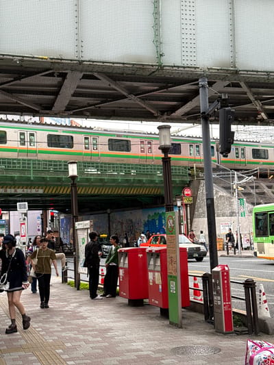 In Shinjuku, Japan, a street scene unfolds beneath a railway overpass. A green and silver train passes overhead on the elevated tracks. Below, a sidewalk is bustling with pedestrians, some walking, others standing and talking. A bright red postal box and a smaller silver fire extinguisher cabinet stand near the sidewalk's edge. Cars and a green bus are visible on the road beyond the sidewalk. The environment is urban, with concrete structures, traffic signals, and various signs indicating local information. The overall atmosphere appears to be a typical day in a busy Japanese city.