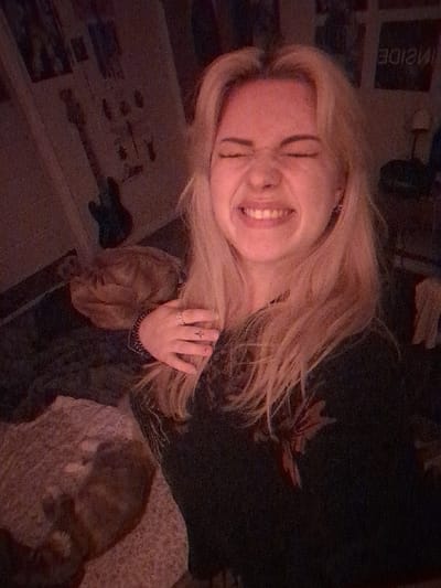 This image depicts a young adult female indoors, located in Area D (Elaho/Garibaldi), Canada. The female, with blonde hair, occupies the central foreground, wearing a dark top with a subtle floral pattern. Her left hand is visible, featuring a ring on her ring finger and a small tattoo, resting near her chest. Her facial expression is characterized by tightly closed eyes, a crinkled nose, and a wide smile revealing teeth, indicating a strong, animated emotion. In the lower-left quadrant, two domestic cats are partially visible, lying on a light-colored, textured surface. The background includes a wall on the left adorned with multiple framed items, one resembling a guitar. On the right, a poster with the word "INSIDE" (appearing reversed) and a small lamp are discernible. The scene is illuminated with a dim, warm, reddish light, contributing to noticeable image grain.