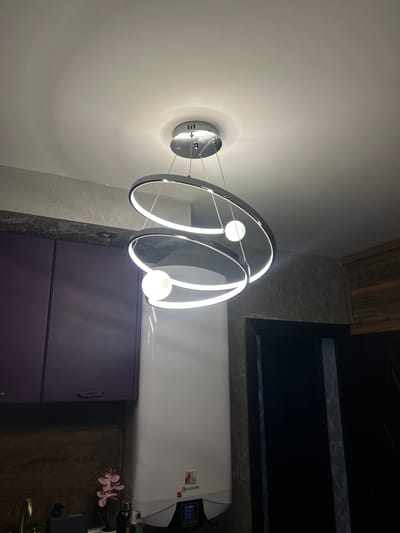 The image depicts a modern living room, possibly in Vidin, Bulgaria, featuring a striking contemporary chandelier with interconnected rings and glowing orbs. The chandelier hangs from a white ceiling, casting a soft light. Below, a wall is textured with a greyish, uneven finish. To the left, a set of sleek, purple cabinets is visible, with a wooden cabinet below them. A white water heater with a digital display is situated on the right. A small pink orchid in a pot adds a touch of natural decor. The overall impression is of a stylish and well-lit interior.