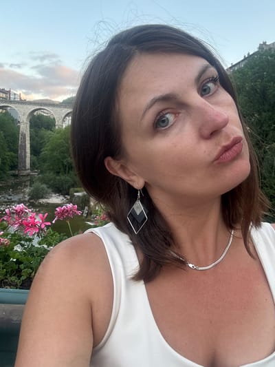 A woman is taking a selfie outdoors with a city bridge in the background. She is positioned on the right side of the frame, her face turned towards the camera. She has brown hair, blue eyes, and is wearing a white top, a silver necklace, and diamond-shaped earrings. In the background, a stone bridge with multiple arches spans over a body of water. Green trees and buildings are visible behind the bridge, and pink flowers are in the foreground. The image appears to have been taken in Saint-Claude, France, as indicated by the presence of the viaduct of the same name.