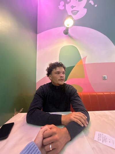 The image shows a man sitting at a table in a restaurant. He is wearing a dark sweater, and his hands are clasped together. Another hand is visible, holding his hand. A receipt sits on the table. A black smartphone is also present. The restaurant features colorful decor on the walls. The image was taken in Serris, France. A light fixture hangs from the ceiling. An electrical outlet is visible on the wall.