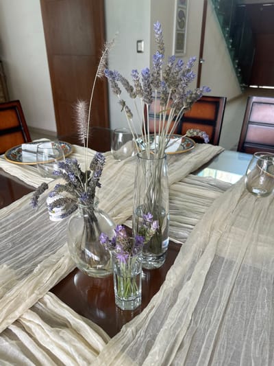 The image shows a table setting indoors. Three transparent glass vases are placed on a beige, draped cloth. Each vase holds sprigs of lavender. One vase also contains feathery grasses. To the left of the vases, there are small plates with a blue and gold rim. The table surface is dark brown wood. In the background, reflective surfaces suggest the interior of a home with stairs and architectural details. The location in Barrio de Nuevo León, Mexico is not verifiable from the content of the image.