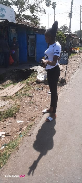 This image depicts a street scene in Homa Bay, Kenya, under clear daylight conditions. In the foreground, a young adult female, facing right in profile, stands on a grey paved surface. She wears a white short-sleeved t-shirt with a dark emblem, black leggings, and white slide sandals. Her hair is styled in upward-braided strands. She holds a dark-colored mobile phone with both hands at abdomen height. Her shadow on the pavement distinctly displays a two-finger "peace sign" hand gesture.
To the immediate left of the paved area is a dirt strip containing sparse green vegetation, dry leaves, and scattered refuse, including pieces of wood and plastic. Beyond this strip stands a small, single-story commercial structure with a corrugated metal roof and blue-painted walls. A partial white sign above its entrance reads "ER'S SHOP". An open doorway reveals a white refrigeration unit inside, and a second individual, wearing a dark top, is partially visible within the shop's interior.
To the right of the standing female, a black A-frame sign with white lettering is partially visible, displaying text such as "SALON" and words indicating operating "DAYS". The background features various other structures, green trees and shrubs, several tall palm trees, and utility poles against a bright sky.