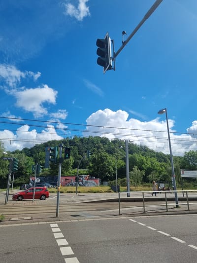 The image shows a street scene in Saarbrücken, Germany, on a bright, sunny day with a blue sky and scattered white clouds. In the foreground, there is an asphalt road with white dashed lines indicating a crosswalk. A red car is visible driving on the road. Across the street, there are tram tracks and a pedestrian area with a few people walking. A traffic light with a green bicycle signal is prominent. Behind the street, a densely wooded hill rises, with some buildings and structures partially visible amongst the trees. Overhead, power lines stretch across the scene, supported by poles. A traffic light hangs suspended from a long arm over the intersection. A street sign that reads "S RÖMERKASTELL" is visible on the right side of the image. The overall impression is of a typical urban environment with a focus on transportation infrastructure and green surroundings.