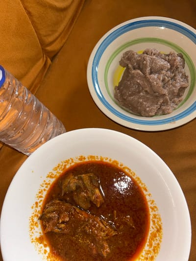 In Egbe, Nigeria, a meal is displayed on a brown, upholstered surface. In the foreground, a white bowl contains a red-orange stew with several pieces of what appears to be meat or fish. A clear plastic water bottle, partially filled with water and featuring a blue cap, rests to the left of the stew bowl. In the background, a second white bowl, adorned with blue and green stripes, holds a quantity of brownish-grey, dense solid food. No individuals are visible in the frame.