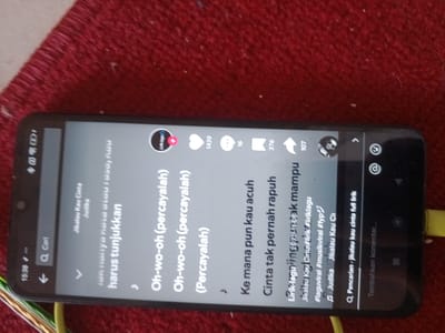 The scene depicts a black smartphone, horizontally oriented and rotated approximately 90 degrees clockwise, resting on a textured, red fabric surface, located in the city of Neuheun, Indonesia. The phone's screen is illuminated, displaying an application interface. In the top left corner of the screen, the time "15:36" is visible, alongside network signal indicators and a battery icon. A search bar labeled "Q Cari" (Search) is present below the status bar.
The central part of the screen displays song lyrics in white text against a dark grey background. Visible lyrics include "harus tunjukkan", "Oh-wo-oh (percayalah)", "Ke mana pun kau acuh", and "Cinta tak pernah rapuh". Above these lyrics, "Jikalau Kau Cinta" and "Judika" are displayed, indicating the song title and artist. To the right of the lyrics, a circular user profile icon, partially visible with text like "Link.Lagu", has a small red plus sign overlay. Below this, interaction metrics are displayed: a heart icon with "1429", a comment bubble icon with "16", a save icon resembling a bookmark with "376", and a share icon with "107". Further text below the main lyrics includes "Lirik-lagu ini puncak mampu", followed by hashtags such as "#laguviral", "#musikviral", and "#fyp", and "Judika Jikalau Kau Ci". The bottom of the screen features a search bar with a magnifying glass icon and "Pencarian - jikalau kau cinta full lirik", next to an input field labeled "Tambahkan komentar..." (Add comment...).
The smartphone has a black casing. A yellow charging cable is connected to the device's port on what would be its bottom edge if held vertically, extending downwards and slightly to the left. The red fabric surface in the background appears to be a carpet or rug, showing visible fibers and some minor white flecks or dust particles.