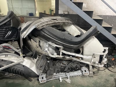 The image shows a pile of car parts, including bumpers, headlights, and structural components, in what appears to be a workshop or storage area. The parts are made of various materials like plastic and metal, and some are damaged or dirty, suggesting they are scrap or from a dismantled vehicle. In the background, there are shelves, stairs, electrical outlets, and some bottles, contributing to the industrial or repair setting. The overall scene is dimly lit, with the car parts being the primary focus. The location is identified as Panyu District, China.