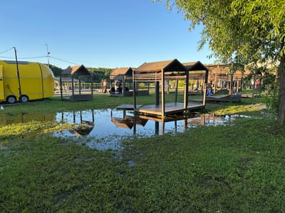 The image depicts an outdoor recreational area, likely a park or beach, with several wooden gazebos situated on platforms. The ground in front of the gazebos is partially flooded, with visible puddles reflecting the structures and the surrounding environment. A bright yellow trailer or food truck is parked on the left side of the image. Several people are scattered around the area, some standing and conversing near the water's edge, while others are seated within the gazebos. The scene is bathed in the warm light of what appears to be late afternoon or early evening, indicated by the long shadows. The background features a body of water, trees, and a clear blue sky. The location is identified as being in the city of Solnechnogorsk, Russia.