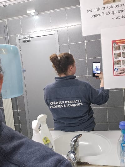 The image depicts a washroom interior, likely within a commercial or public building in Mouscron, Belgium. The scene is captured from a mirror's perspective, showing the back of an individual and elements behind them.

A person, identified as female, stands with her back to the viewer, facing the mirror. She has reddish-brown hair tied in a bun with a small, light-colored clip. She wears a dark blue long-sleeved shirt with white text on the back that reads: "CRÉATEUR D'ESPACES PROPRES & LUMINEUX" (Creator of Clean & Bright Spaces). In her right hand, she holds a black smartphone, pointed towards the mirror, with its screen displaying a reflection of the scene.

In the foreground, reflected in the mirror, are elements of a washbasin area. A white ceramic sink is centered, equipped with a chrome faucet. To the left of the sink, two white spray bottles, likely cleaning products, are visible, one with a partial yellow label. To the right, a clear plastic bottle with a blue cap is present.

The background, also reflected, features walls tiled with dark gray square tiles and white grout. A light-colored metal door with an overhead door closer mechanism is visible on the left. Above the door, a light fixture is mounted. To the left of the person's head, a light blue hand dryer or paper towel dispenser is partially visible. Two white signs are affixed to the tiled wall to the right of the person. The upper sign displays mirrored French text, indicating handwashing instructions. The lower sign is a detailed poster with multiple pictograms illustrating proper hand hygiene steps.