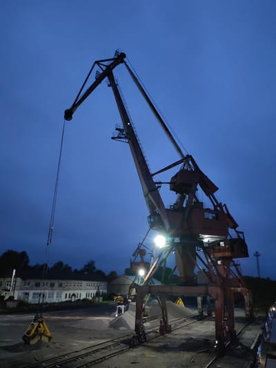 This image captures a massive, dark reddish-brown industrial port crane silhouetted against a deep blue, twilight sky. The crane's intricate structure, including its towering boom and counterweights, extends high into the frame. Bright artificial lights are integrated into the crane's structure, illuminating parts of its body and the ground below.

Hanging from chains connected to the crane's cable is a large, yellow grapple bucket, suspended just above the ground in the foreground. Several parallel rail tracks run across the foreground and beneath the crane's base. To the right, large piles of sand or gravel are visible, brightly lit by the crane's lights. A white truck is partially visible near these material piles, suggesting active work or preparation.

In the background to the left, a long, two-story, light-colored building with multiple windows can be seen, partially obscured by dark trees. The scene depicts an industrial loading or port area, likely in Medvezhyegorsk, Russia, during the early evening or night, indicated by the dark sky and prominent artificial lighting. The weather appears clear and calm. The number "52" is clearly visible on a white plate attached to the main body of the crane. No people are discernible, and no specific activity is actively taking place, though the setup suggests bulk cargo handling operations.