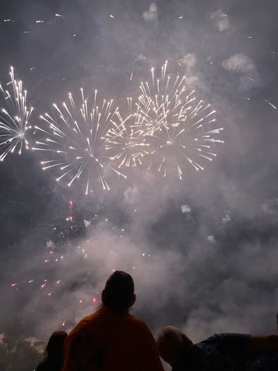 The image captures a nighttime fireworks display over the city of Canet de Mar, Spain. In the foreground, silhouetted figures of people are visible from behind, looking up at the sky. The sky is filled with bursts of white fireworks, creating bright streaks and patterns against a dark, cloudy backdrop. Scattered throughout the sky are smaller, colorful fireworks, appearing as red and green sparks. Wisps of smoke and clouds mingle with the illumination from the fireworks. The scene suggests a festive occasion, likely a celebration or holiday.