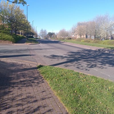 The image shows a street scene in Coventry, United Kingdom. It is a sunny day with a clear blue sky. The street is asphalt and has double yellow lines. There is a pedestrian pavement on the left side of the image, made of red bricks. There is green grass to the right of the pavement. The street is lined with trees on both sides, casting shadows on the road. In the distance, there are buildings and traffic lights. There are some cars far away to the left of the scene.