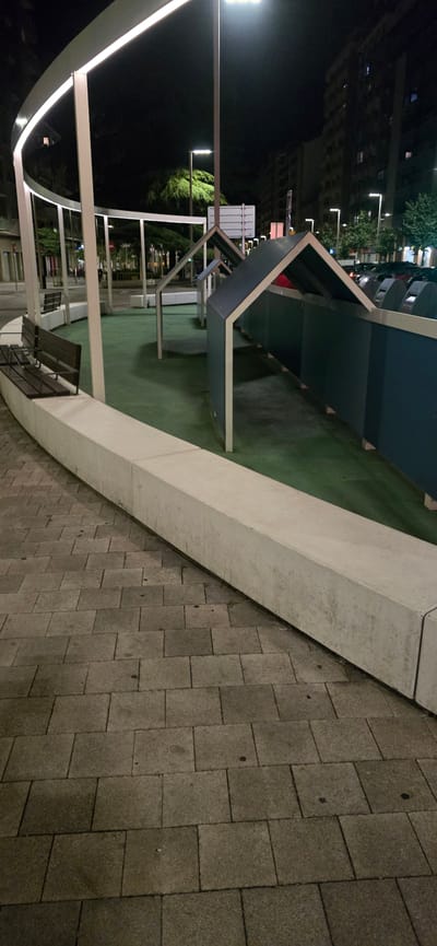 The image captures an outdoor scene at night in Huesca, Spain, featuring a modern architectural installation. The primary subjects are the geometric structure itself, composed of dark blue panels and white metal framing, and the surrounding paved area. The ground within the installation appears to be covered with artificial turf, a vibrant green against the darker surroundings.
The scene is illuminated by artificial lighting, including streetlights and integrated lighting within the architectural elements, which casts distinct shadows and highlights. The time of day is clearly night, with the dark sky and the presence of artificial illumination indicating this. The background shows the faint outlines of buildings and trees, suggesting an urban or semi-urban environment.
Notable details include the texture of the paving stones, which are rectangular and laid in a staggered pattern, and the rough, concrete-like finish of the curved barrier bordering the paved area. The architectural installation has an open, angular design, with multiple planes and supports. There are no people visible in the frame, and the overall mood is one of quiet and perhaps contemplative observation of the urban design. No visible text is present in the image.