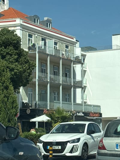 The image depicts a street scene in Oeiras, Portugal, featuring a multi-story building with a red tiled roof and ornate architectural details. The building has balconies with metal railings on several floors, and some of the balconies appear to have outdoor furniture. A large green tree is positioned to the left of the building, and a modern white building is visible to its right. In the foreground, two cars are parked, one black and one white. The white car has a license plate that reads "90 TX". There are signs visible on the facade of the lower part of the building, with neon lettering that appears to say "Calde" and "Pizzaria" or "Ristorante". A white umbrella is set up outside a storefront. A person wearing a green and white garment is partially visible amongst some bushes in front of the building. The sky is clear and blue.