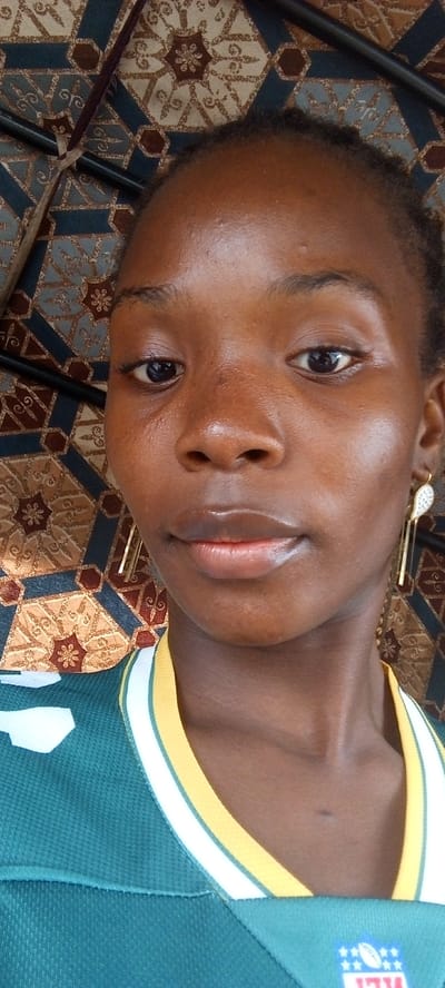 A close-up portrait of a young Black woman with clear skin, dark eyes, and full lips. She is wearing a green and yellow collared shirt with a white stripe. Her earrings are visible. The background is a patterned brown, blue, and gold textile. It is impossible to know for sure if the location is Uke, Nigeria.