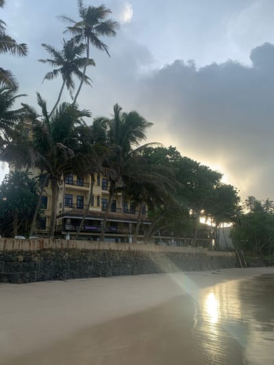 This is a photograph of a beachfront in Kathaluwa, Sri Lanka, at what appears to be either sunrise or sunset, given the low angle of the sun and the reflection on the wet sand. In the background, a multi-story building with a yellow facade and dark windows is visible, framed by numerous tall palm trees. The building has balconies with dark railings and a tiled roof section. A stone retaining wall, reinforced with wire mesh cages filled with rocks, separates the beach from the building's grounds. The foreground is dominated by a sandy beach with the tide line marked by wet sand reflecting the sky. The sky is overcast with dramatic clouds, but a patch of brighter sky suggests the sun is near the horizon, creating a lens flare effect on the water's surface. There are no people or animals visible in the image.