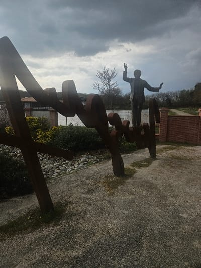 This image depicts a public art installation in Avignonet-Lauragais, France, under an overcast sky. In the foreground, a large, dark, rust-colored abstract metal sculpture, composed of curvilinear and angular forms, extends from the left margin towards the right. This sculpture appears to form a word or name, with its supports rooted in a light-colored gravel path that covers the immediate ground. Behind this abstract sculpture, slightly to the right of the center, stands a full-body statue of a male figure. This statue is dark, likely bronze or metal, depicting a man in a suit jacket and trousers with both arms raised expressively, his right arm bent at the elbow and raised higher than his left.
Separating the statues from the background is a body of water, presumably the Canal du Midi. Along the water's edge, between the foreground sculpture and the male statue, is a strip of green foliage and bushes. Behind the male statue, a small tree with delicate reddish-purple blossoms is visible. To the right of the male statue, a low brick wall section is present. The far bank of the waterway features further green foliage and distant trees. A building with a dark roof and light-colored walls is visible across the water, slightly to the left of the male statue. The sky above is largely covered with thick, grey clouds, and the overall lighting is soft, indicative of an overcast day.