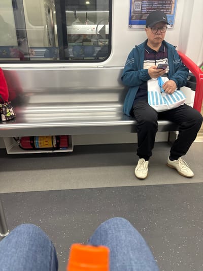 Inside a train car, an older man with a baseball cap and glasses is seated, holding a phone and a striped bag. He wears a teal jacket, patterned shirt, and black pants, with off-white sneakers. An adjacent passenger's jean-clad legs are visible. The train interior features a silver wall, a window revealing outside scenes, and an extinguisher. The floor has a speckled gray pattern.