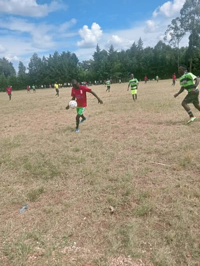 An outdoor football match is underway on a dry, grassy field in Lwandeti ward, Kenya. Approximately 15-20 male individuals, primarily young adults, are visible across the scene. The central figure, wearing a red short-sleeved jersey with white text and green shorts, is actively dribbling a white and black football with his right foot, positioned mid-stride. To his right, another player in a green and black striped jersey with black shorts is moving towards him. Other players, dressed in either red jerseys and green shorts or green/black striped jerseys and black shorts, are dispersed across the field. A line of tall trees forms the background. The sky is blue with scattered white clouds. The ground consists of brown, dry grass with patches of green and some visible small rocks. A clear plastic bottle lies discarded in the lower-left foreground.