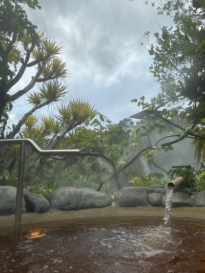 The image shows a hot spring in Ban Khlong Khian, Thailand. Water flows from a bamboo pipe into a stone-lined pool. Lush vegetation surrounds the pool, including trees with spiky leaves. A metal handrail is visible. Steam rises from the water, creating a misty atmosphere. The sky is cloudy.