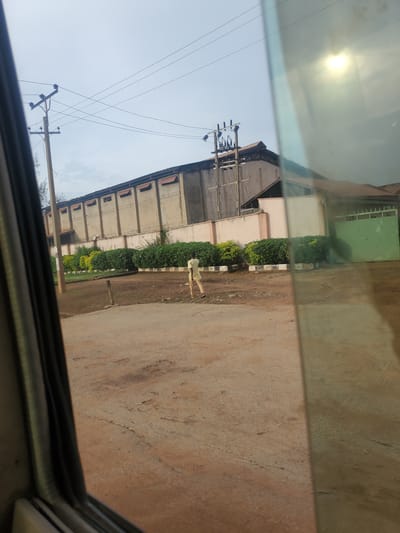 This is a medium-shot, eye-level, outdoor, daytime, vertical, medium-contrast, slightly blurry, color photograph of a person walking near a large, utilitarian building. The scene appears to be located in Television, Nigeria.
In the foreground, a dirt road occupies the bottom half of the image, showing cracks and inconsistencies in the surface. A portion of a dark-colored vehicle's interior is visible along the left edge, framing the scene.
In the mid-ground, a lone individual, appearing to be an adult male wearing a light-colored shirt and pants, is walking away from the camera towards the right. He is positioned in front of a line of manicured green bushes. Beyond the bushes, a low, light-colored wall with decorative elements and barbed wire on top separates the grounds from the building.
The background is dominated by a large, multi-story building with a light-colored, textured facade and a sloped, dark roof. The building has a series of regularly spaced openings along its upper section. Electrical poles and wires are visible in front of and above the building, indicating an industrial or commercial area. The sky is overcast with some bright patches, suggesting diffused sunlight. A green gate is visible on the far right of the building. The overall impression is one of an industrial or factory setting.