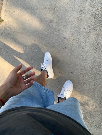 The image is a first-person perspective shot of a person standing on a concrete surface outdoors. The person is wearing white sneakers, denim shorts, and a dark t-shirt. Their left hand, adorned with a ring, is extended towards the camera. Sunlight casts shadows across the concrete, which has a textured, wavy pattern. There is no visible indication that this scene is located in El Valle del Espíritu Santo, Venezuela.