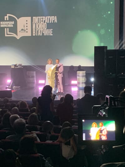 The image depicts an indoor event, likely a film festival or cultural gathering, as suggested by the text "ЛИТЕРАТУРА И КИНО В ГАТЧИНЕ" (Literature and Cinema in Gatchina) and "МЕЖДУНАРОДНЫЙ КИНОФЕСТИВАЛЬ" (International Film Festival) visible on a large screen in the background. The scene appears to be a stage with several people on it, and an audience seated in rows of chairs.
On the stage, three individuals are prominent. Two are standing side-by-side, one wearing a light pink suit and the other a light yellow and white outfit. Between them, or slightly in front, is a golden, somewhat abstract object that resembles a stylized feather or leaf. The third person on stage is partially obscured. The stage is lit with bright spotlights, creating a dramatic effect, and there are several rows of stacked horizontal elements behind the people, possibly stage props or sound equipment. A monitor screen is visible in the upper right corner, displaying a magnified view of the people on stage.
The audience members are mostly silhouettes, with their faces turned towards the stage. The overall mood seems to be one of attention and engagement, with people focused on the proceedings. The time of day is not explicitly indicated, but the indoor lighting suggests it could be evening or night. The event is taking place in Gatchina, Russia, as confirmed by the text.
