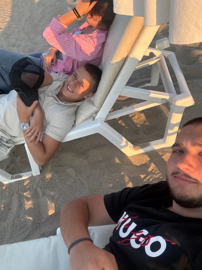 Three individuals are relaxing on a sandy beach in Sveti Vlas, Bulgaria, near white lounge chairs. The person in the foreground is taking a selfie, wearing a black t-shirt with a "HUGO" logo. To their left, a young man is lying on a lounge chair, holding a black baseball cap and wearing a light-colored polo shirt and a watch. Behind him, another person is partially visible, wearing a pink and white striped shirt and blue jeans. The sun is casting long shadows on the sand, indicating it may be late afternoon or early evening.