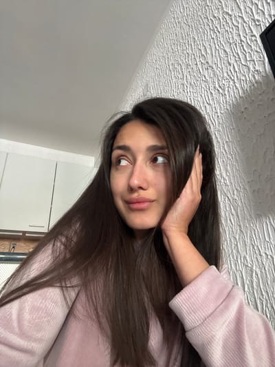 A young woman with long, dark hair is holding her hand to her head. She is wearing a pink sweatshirt. The background includes a textured ceiling and a gray wall with white cabinets. The location is not specified or discernible from the image.